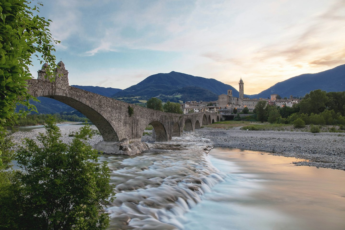 Il Ponte Gobbo in pietra attraversa un fiume roccioso vicino a un paese.